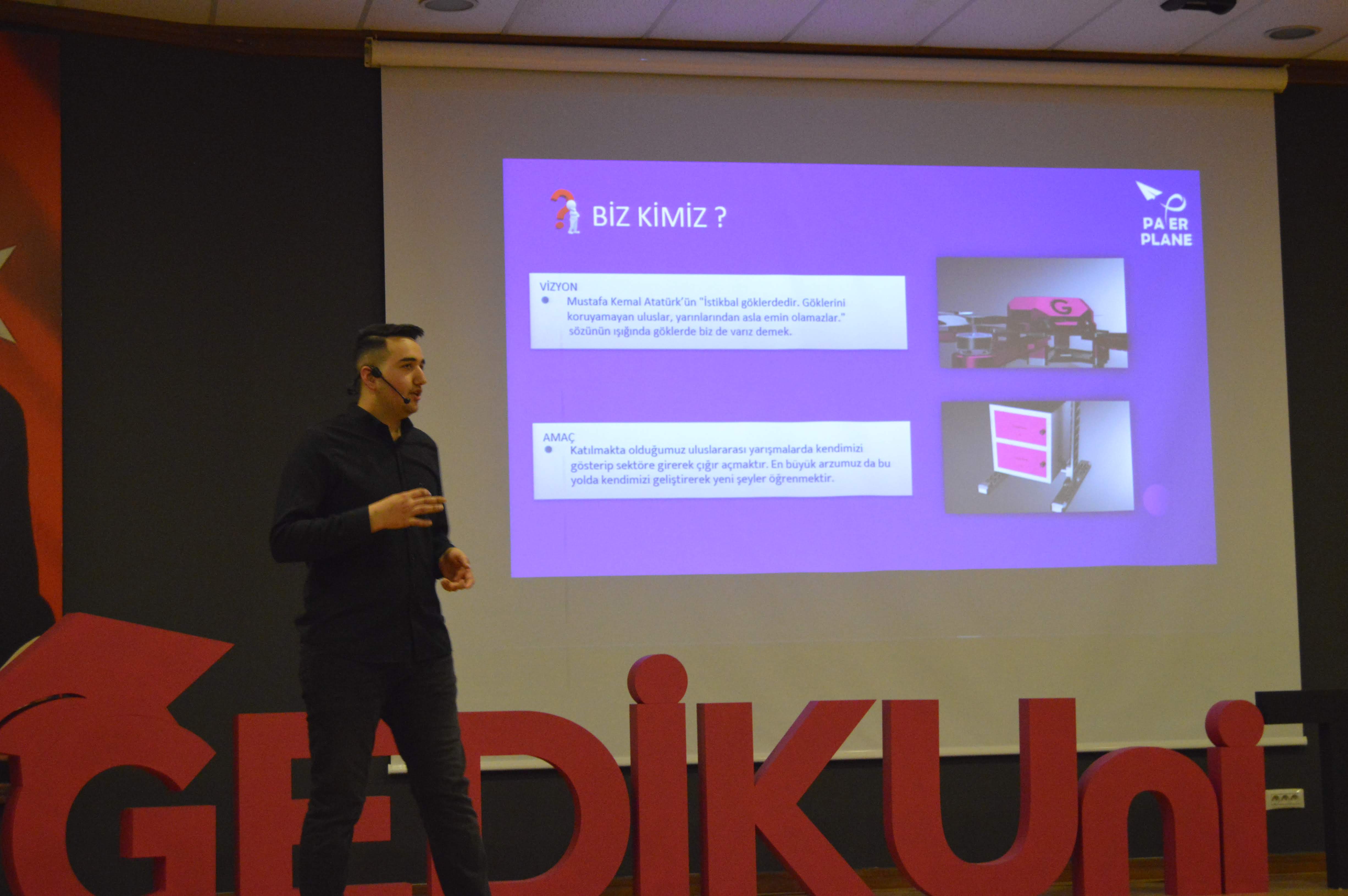 Mehmet Seyrimez, İstanbul Gedik Üniversitesi sahnesinde Paper Plane İHA takımını tanıtan “Biz Kimiz?” slaytı önünde sunum yaparken